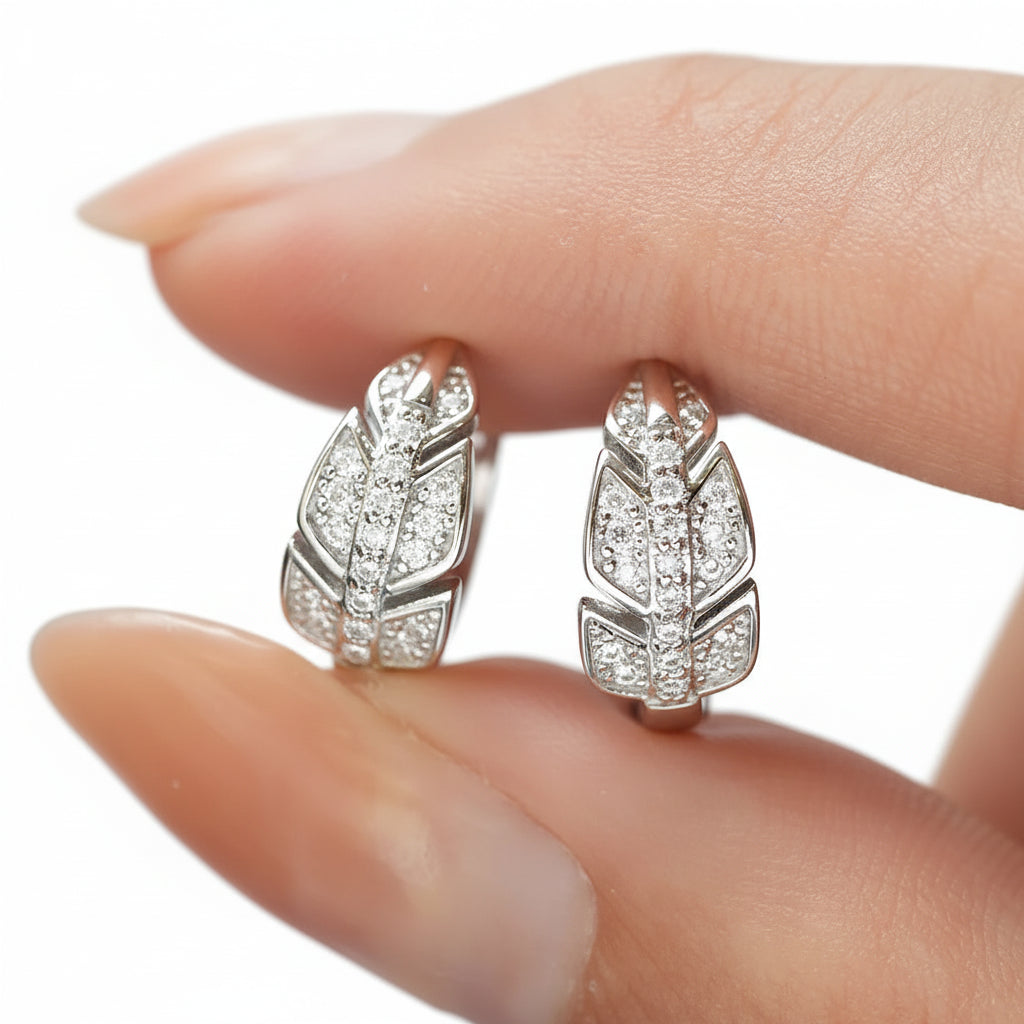 Silver feather-shaped earrings with gemstones held between fingers against a blurred background